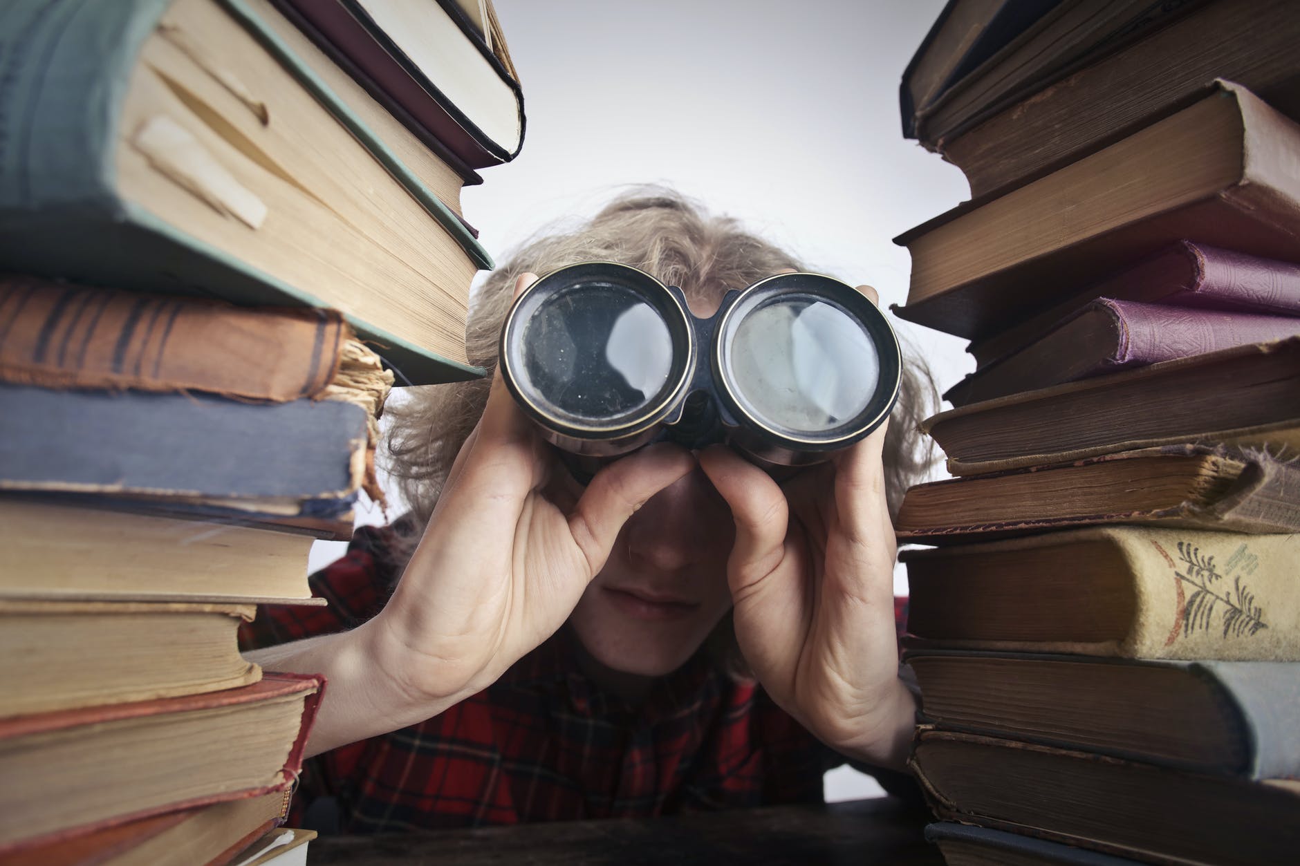 person with blond hair looking through binoculars between 2 stacks of old books